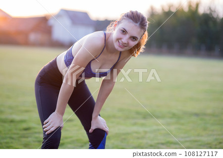 Slim blonde girl plays sports and performs yoga poses in summer grass covered stadium on a sunset background. Woman doing exercises on the yoga mat. 108127117
