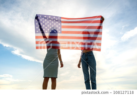 Silhouette of two young friends women holding USA national flag in their hands at sunset. Patriotic girls celebrating United States independence day. International day of democracy concept. 108127118