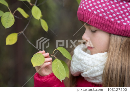 Pretty child girl wearing warm winter clothes holding tree branch with green leaves in cold weather outdoors. Pretty child girl wearing warm winter clothes holding tree branch with green leaves in cold weather outdoors. 108127200