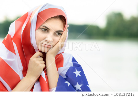 Portrait of young sad refugee woman with USA national flag on her head and shoulders. Immigration in United States concept. 108127212