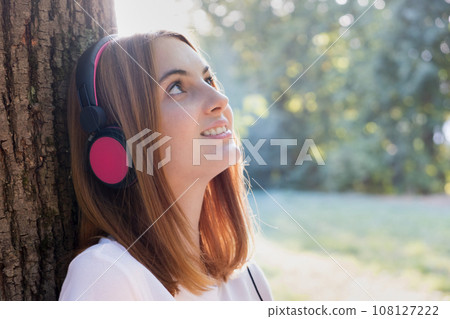 Portrait of red haired teenage girl listening to music in big pink earphones outdoors leaning to a tree. Portrait of red haired teenage girl listening to music in big pink earphones outdoors leaning to a tree. 108127222