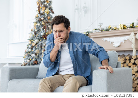 A young man sits at home on the couch during the New Year holidays and feels very nauseous, covers his mouth with his hand, wants to vomit. Alcohol intoxication, poisoning. A young man sits at home on the couch during the New Year holidays and feels very nauseous, covers his mouth with his hand, wants to vomit. Alcohol intoxication, poisoning. 108127395