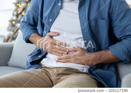 Close-up photo of young man's hands holding his stomach. A man sits on a couch and suffers from pain, spasms, poisoning, overeating. 108127396