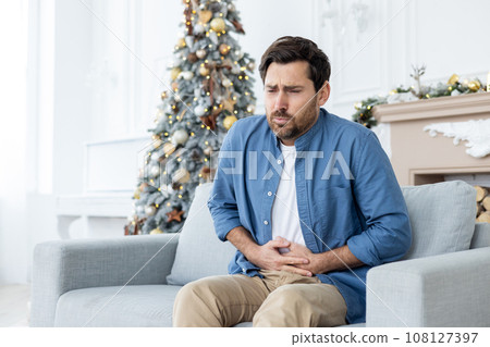 A young man suffers from severe abdominal pain during the Christmas holidays. He sits at home on the sofa near the Christmas tree and holds his body with his hands, grimacing from spasms. A young man suffers from severe abdominal pain during the Christmas holidays. He sits at home on the sofa near the Christmas tree and holds his body with his hands, grimacing from spasms. 108127397