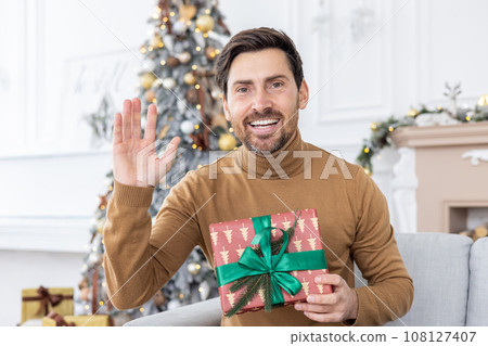 Portrait of a smiling young man talking to the camera via video link. Sitting at home on the sofa near the Christmas tree and holding a New Year's gift in his hands, greeting and waving. 108127407