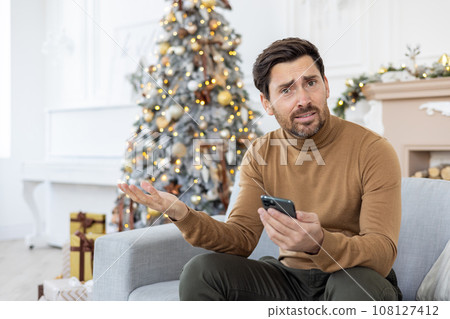 Portrait of a young man sitting at home on New Year's holidays near the Christmas tree on the sofa, using the phone and unhappily waving his hands at the camera. Portrait of a young man sitting at home on New Year's holidays near the Christmas tree on the sofa, using the phone and unhappily waving his hands at the camera. 108127412