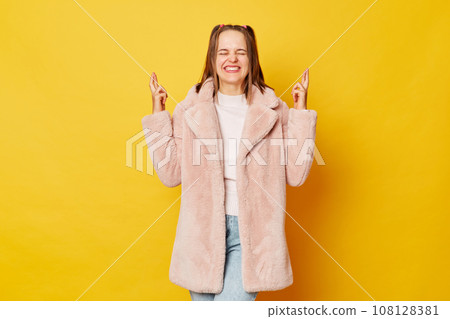 Smiling young girl with funny ponytails standing isolated over yellow background holding fingers crossed for good luck having desire closed eyes. Smiling young girl with funny ponytails standing isolated over yellow background holding fingers crossed for good luck having desire closed eyes. 108128381