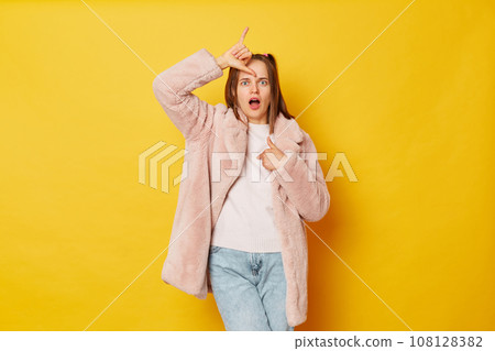 Upset disappointed woman wearing fur coat standing, showing looser gesture with hand on forehead and pointing herself looking at camera isolated over yellow background. Upset disappointed woman wearing fur coat standing, showing looser gesture with hand on forehead and pointing herself looking at camera isolated over yellow background. 108128382