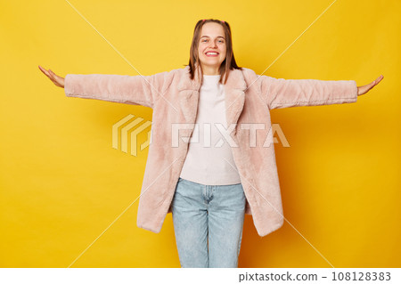 Charming adorable woman dressed pink fur coat isolated over yellow background spreading arms pretending flying looking smiling at camera expressing happiness. Charming adorable woman dressed pink fur coat isolated over yellow background spreading arms pretending flying looking smiling at camera expressing happiness. 108128383