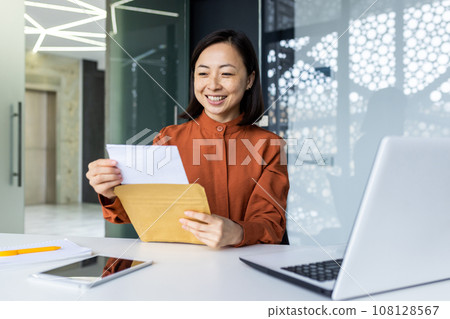 Successful satisfied business woman received mail envelope letter with good news message, Asian woman at workplace reading document paper, smiling at workplace inside office. 108128567