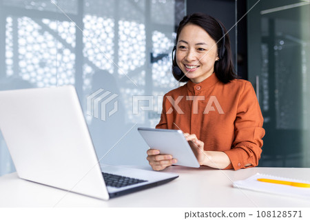 Young successful asian woman inside office at workplace with tablet computer and headset phone, woman advises buyers consumers remotely, business woman use laptop for video call. 108128571