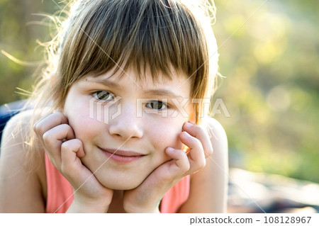 Portrait of pretty child girl with gray eyes and long fair hair leaning on her hands smiling happily outdoors on blurred bright background. Cute female kid on warm summer day outside. Portrait of pretty child girl with gray eyes and long fair hair leaning on her hands smiling happily outdoors on blurred bright background. Cute female kid on warm summer day outside. 108128967