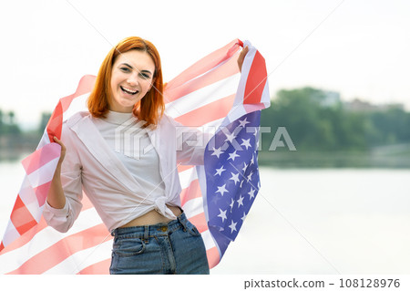 Portrait of happy smiling red haired girl with USA national flag on her shoulders. Positive young woman celebrating United States independence day. 108128976