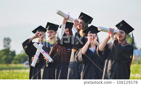 Cheerful graduates on a sunny day look through diplomas like a telescope. 108129178