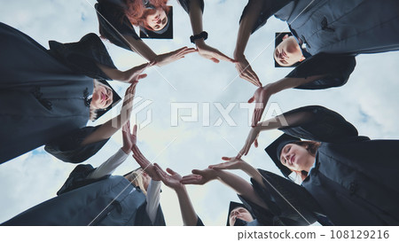 Graduate students in black robes and caps make a circle shape from their hands. 108129216