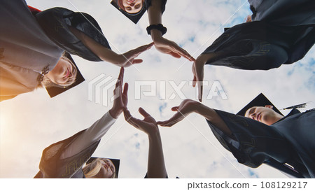 Graduate students in black robes and caps make a circle shape from their hands. 108129217