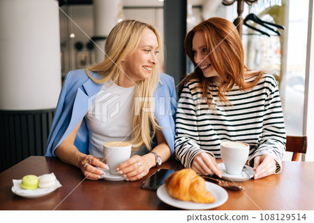 Portrait of cheerful young best girlfriends sitting at modern cafe, communicating and sharing stories, spending relax in coffee shop. Two women friends sitting in cafeteria and talking. Portrait of cheerful young best girlfriends sitting at modern cafe, communicating and sharing stories, spending relax in coffee shop. Two women friends sitting in cafeteria and talking. 108129514