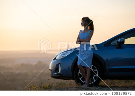 Happy woman driver in blue summer dress enjoying warm evening near her car. Travel and vacations concept. Happy woman driver in blue summer dress enjoying warm evening near her car. Travel and vacations concept. 108129872