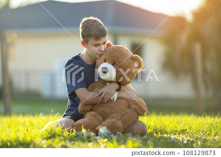 Happy teenager boy hugging his teddy bear friend outdoors on green grass lawn. Friendship concept Happy teenager boy hugging his teddy bear friend outdoors on green grass lawn. Friendship concept 108129873