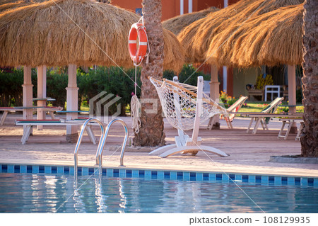 Empty deck chairs under straw shade umbrellas on swimming pool side in tropical resort. Summer vacations and getaway concept Empty deck chairs under straw shade umbrellas on swimming pool side in tropical resort. Summer vacations and getaway concept 108129935