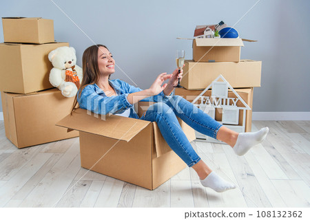 Happy woman drinks champagne from a glass while sitting inside a cardboard box in new apartment. 108132362