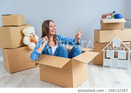 Happy woman drinks champagne from a glass while sitting inside a cardboard box in new apartment. Happy woman drinks champagne from a glass while sitting inside a cardboard box in new apartment. 108132367