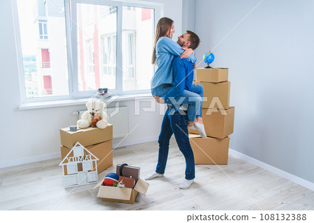 Front view of pretty smiling girl which holds by her handsome young boyfriend in the middle of their new flat full of cardboard boxes. Front view of pretty smiling girl which holds by her handsome young boyfriend in the middle of their new flat full of cardboard boxes. 108132388