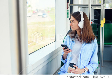 Young woman using public transport sits with smart phone and white headphones in the modern bus. Young woman using public transport sits with smart phone and white headphones in the modern bus. 108132395