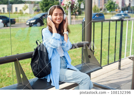 Teen girl listens to the music by white headphones in a public transport station while she waiting for tram. Teen girl listens to the music by white headphones in a public transport station while she waiting for tram. 108132396