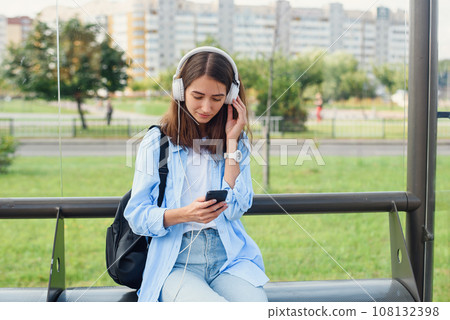 Teen girl listens to the music by white headphones in a public transport station while she waiting for tram. Teen girl listens to the music by white headphones in a public transport station while she waiting for tram. 108132398