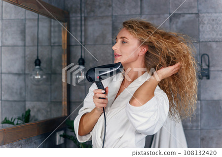 Charming smiling lady with light curly hair in white dressing gown looking at her reflection in the bathroom mirror and drying her hair with hair drayer Charming smiling lady with light curly hair in white dressing gown looking at her reflection in the bathroom mirror and drying her hair with hair drayer 108132420