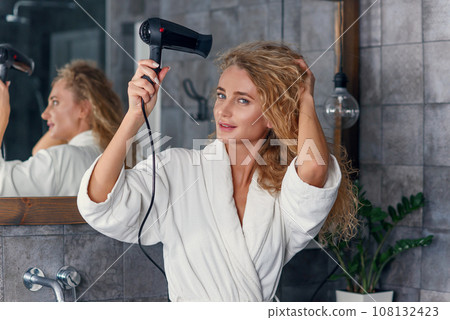 Close up of appealing smiling young lady in white dressing gown posing on camera while drying her curly hair using hair dryer near the bathroom mirror 108132423