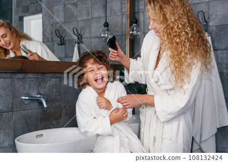 Charming woman dressed in white bathrobe combs her son's curly hair while he sitting on washbasin and laughing. 108132424