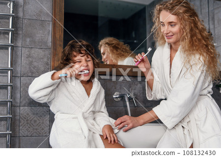 Cheerful little boy with curly hair in a white bathrobe brushes his teeth with toothpaste and a toothbrush with his pretty mom in bathroom at the morning. 108132430