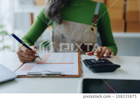 Small business, online business owner, young woman is using profit calculator and checking inventory. 108135875