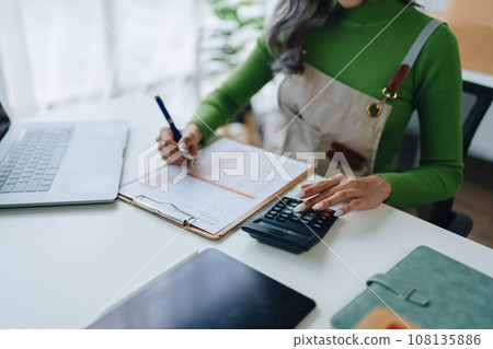 Small business, online business owner, young woman is using profit calculator and checking inventory. Small business, online business owner, young woman is using profit calculator and checking inventory. 108135886