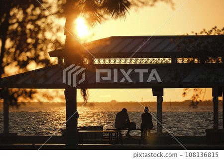 Dark silhouette of people resting under alcove roof on sea shore in public park at sunset Dark silhouette of people resting under alcove roof on sea shore in public park at sunset 108136815