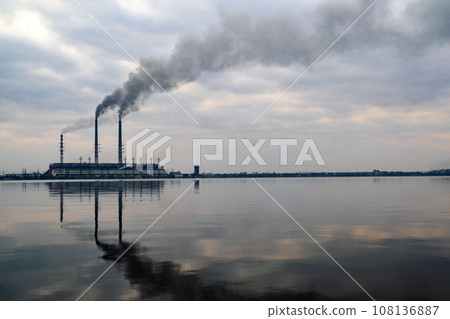 Coal power plant high pipes with black smoke moving upwards polluting atmosphere with reflections of it in lake water. Coal power plant high pipes with black smoke moving upwards polluting atmosphere with reflections of it in lake water. 108136887