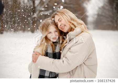Mother and daughter having fun in forest on snow day. Woman wearing beige warm jacket and girl wrapping in a plaid blanket. Woman and teen girl hugging. Mother and daughter having fun in forest on snow day. Woman wearing beige warm jacket and girl wrapping in a plaid blanket. Woman and teen girl hugging. 108137426