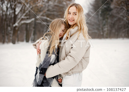 Mother and daughter having fun in forest on snow day. Woman wearing beige warm jacket and girl wrapping in a plaid blanket. Woman and teen girl hugging. Mother and daughter having fun in forest on snow day. Woman wearing beige warm jacket and girl wrapping in a plaid blanket. Woman and teen girl hugging. 108137427