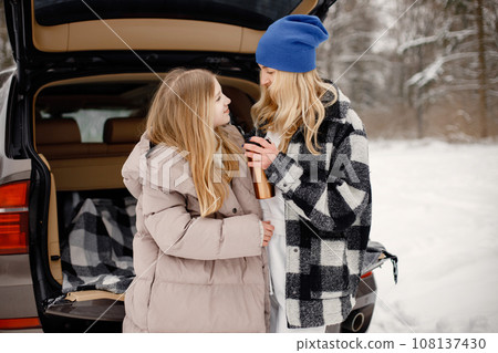 Mother and daughter having fun in forest on snow day. Woman and girl wearing beige warm jackets. Woman and teen girl standing near open car's trunk and holding a thermos with tea. 108137430