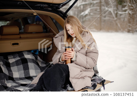 Young blonde girl sitting in an open car's trunk. Girl holding thermos with tea in hands. Teenager wearing beige jacket. Young blonde girl sitting in an open car's trunk. Girl holding thermos with tea in hands. Teenager wearing beige jacket. 108137451
