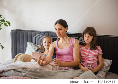 Mother with two daughters lying on a bed under blanket. fFamily making video call to father. Concept of Mother's Day and maternal love. 108139641