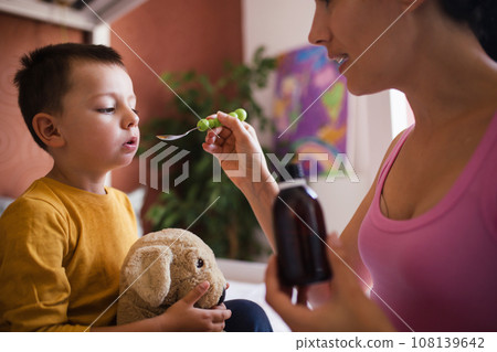 Young mother giving medicines, coughing syrup from spoon to her sick little son. Concept of children seasonal illness, viruses and kid's immune system. 108139642