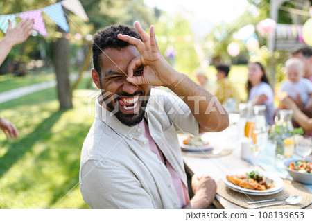 Handsome man making funny faces, sitting at the party table during a summer garden party with family and friends. Handsome man making funny faces, sitting at the party table during a summer garden party with family and friends. 108139653