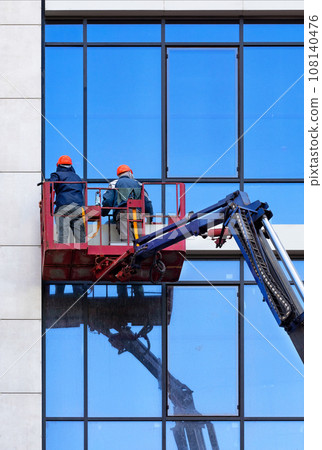 Two men cleaning windows of an exterior glass facade on a construction lifting cradle clean the glass facade of a building on a clear day against the backdrop of a blue reflected sky. 108140476