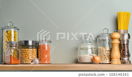 Various uncooked groceries and pasta in glass jars arranged on wooden shelf on olive green wall. Various uncooked groceries and pasta in glass jars arranged on wooden shelf on olive green wall. 108148161