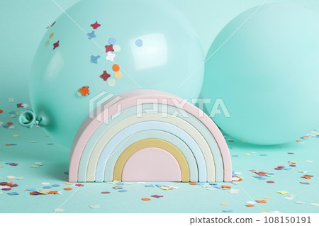 a plastic rainbow toy in front of balloons of the same color as the background and confetti on the ground. 108150191