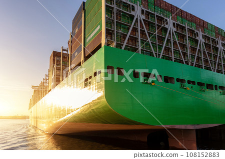 Scenic view giant cargo container ship loading Hamburg city port harbour against seaport cranes warm evening sunset lights. Global commercial trade freight charter shipping and logistics background Scenic view giant cargo container ship loading Hamburg city port harbour against seaport cranes warm evening sunset lights. Global commercial trade freight charter shipping and logistics background 108152883