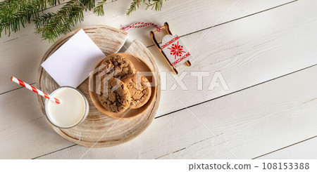 Top view Christmas composition. Glass milk, cookies, Santa's sleigh, empty note, white wooden table. 108153388